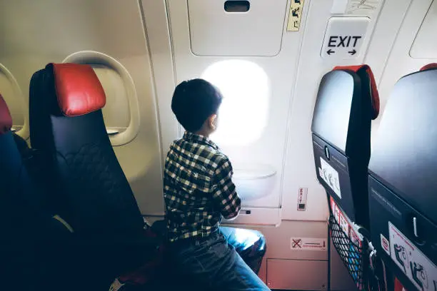 Rear view of a young boy flying on his flight and staring out the window of an airplane.