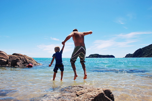 A man holds hands with a young boy whilst they leap off the rocks into the sea.