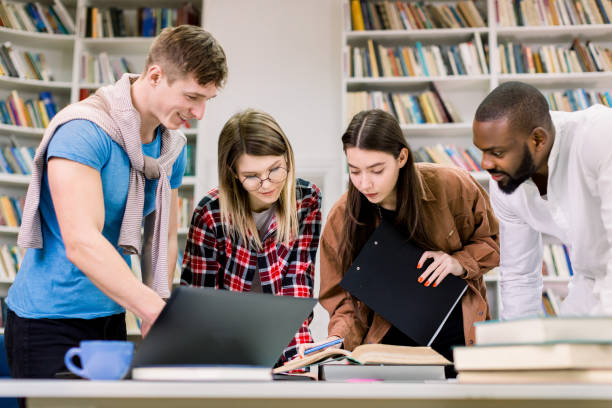 istock Multiethnical attractive smiling team of students, coworkers which are working and communicating together over home task, exam or project in the library, using books and laptop 1219859942
