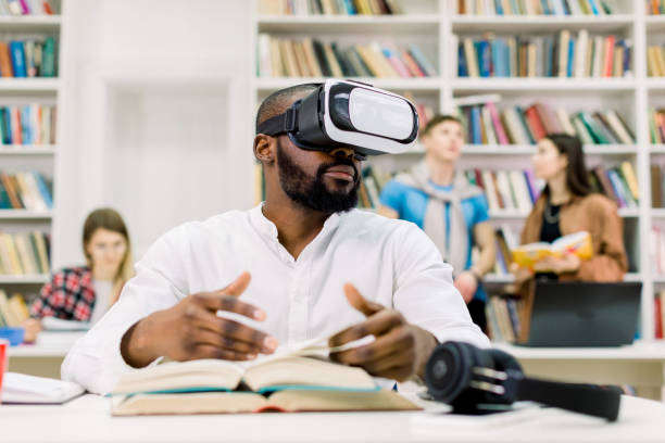 istock Young attractive African man with beard wearing casual white shirt, studying in library, using virtual reality goggles headset, flipping through book pages. Diverse students studying on the background 1219860504