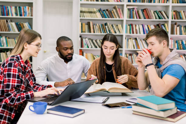 istock Front view of four good-looking modern smart mixed race students which are sitting in the reading room of library and preparing for exams or scientific project. Books, laptop, tablet on the table 1219860678