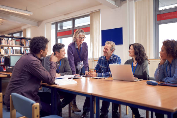 teacher with group of mature adult students in class sit around table and work in college library - night school imagens e fotografias de stock