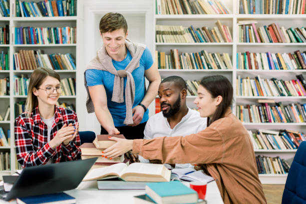 istock Handsome satisfied young man student putting many different books on the table for his university multi racial friends, sitting and studying together in modern campus library reading room. 1219860825