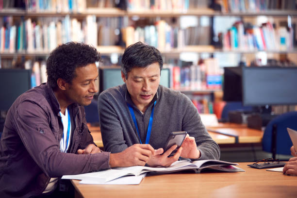 teacher with mature male adult student using mobile phone at table working in college library - night school imagens e fotografias de stock