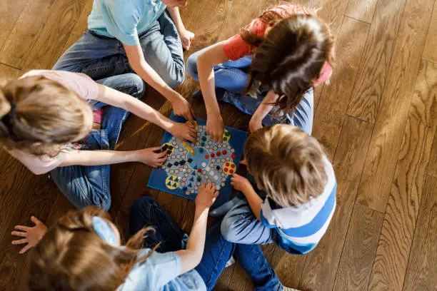 High angle view of group of small friends playing ludo game on wooden floor at home.