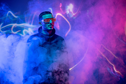 Serious cool guy in neon goggles and sweatshirt standing in blue and pink smoked room