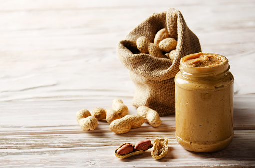 Low angle view at glass mason jar with peanut butter on white wooden table with burlap sack on backside. Healthy eating concept