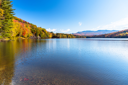 Beautiful mountain lake surrounded by colourful deciduous trees at the peak of fall foliage on a sunny morning. Reflection in water. Waterbury, VT, USA.