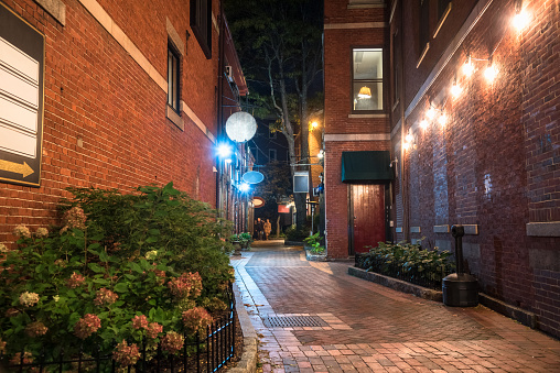 Alley lined with old brick buildings with restaurants on the ground level at night. Pepeople walking along the alley are visible in distance. Portsmouth, NH, USA.