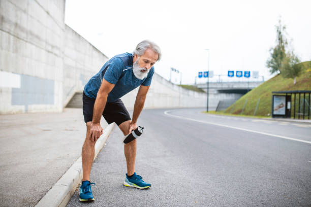 Exhausted Mature Male Runner Stopping for Rest and Water Bearded Caucasian runner in mid 50s holding water bottle and bending over with hands on knees as he rests from daily exercise on deserted urban street. hand on knee stock pictures, royalty-free photos & images