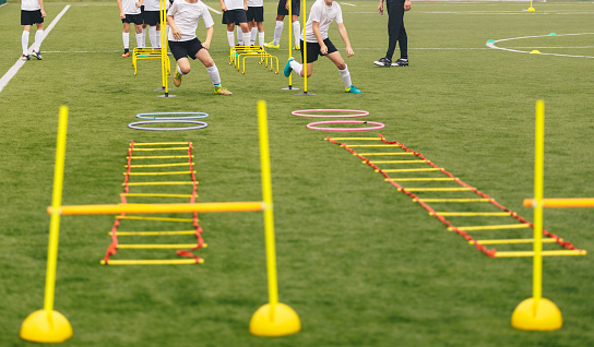 Soccer field with training equipment and players with coach in background. Junior football team training and coach. Sport team on training. Football training equipment: ladder, hula hoop, hurdle, cone