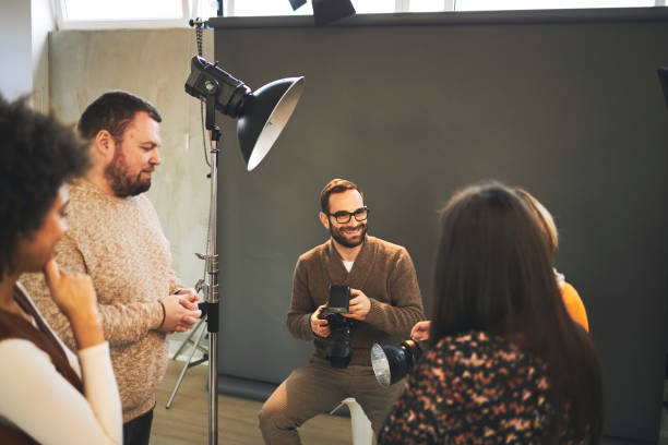 istock Young attractive bearded tutor sitting on chair in studio, holding camera and explaining something about photography while attendees standing and looking at him. 1219879418