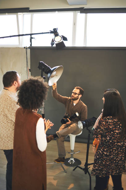 istock Young attractive bearded tutor sitting on chair in studio, holding camera and explaining something about photography while attendees standing and looking at him. 1219879501