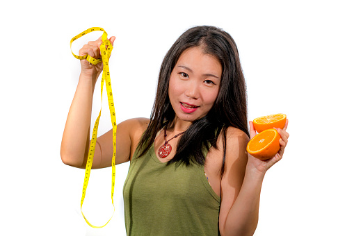 young happy and beautiful Asian Korean woman holding orange fruit and tape measure smiling cheerful in diet and weight loss through healthy natural nutrition isolated on white