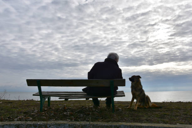 Senior man sitting on an old wooden bench by the sea with his dog quietly standing by. Dark cloudy winter day. Nostalgic mood.
