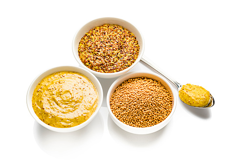 Spices: two bowls filled with whole grain and Dijon mustard shot on white background. The third bowl is filled with dried mustard seeds. A spoon with mustard is beside the bowls. Predominant colors are gold and white. High resolution 42Mp studio digital capture taken with SONY A7rII and Zeiss Batis 40mm F2.0 CF lens