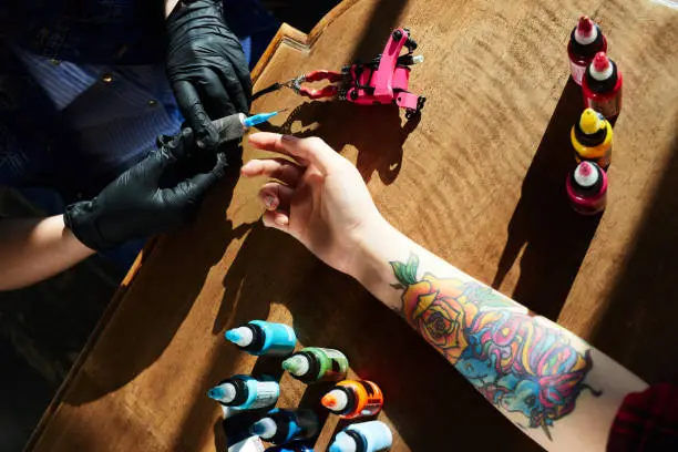 Tattoo expert in gloves holding tattoo gun while hand of female client with new colorful tattoo lying in wooden table beside, high angle close-up view