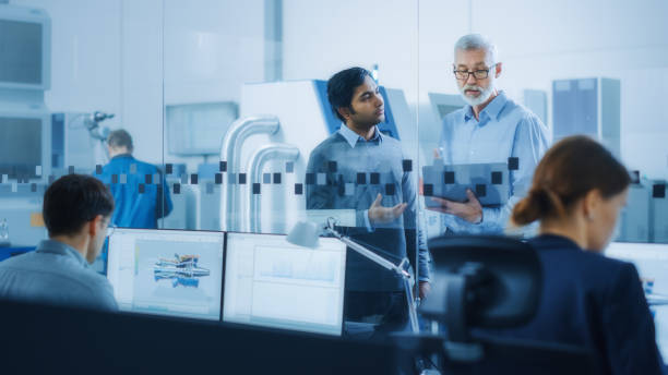 istock Automotive Design Factory: Project Manager, Chief Mechanical Engineer Talk, Use Laptop, Discuss Project Details. High-Tech Industrial Facility Construction of Engines using CNC Machinery. 1219894317