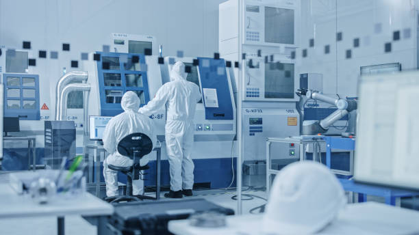 sterile moderne fabrik: profis in coveralls, masken arbeiten auf cnc-maschinen. medizinisches elektronik-fertigungslabor mit high-tech-roboterarm-produktionslinie und zeitgenössischer ausrüstung - elektrotherapie stock-fotos und bilder