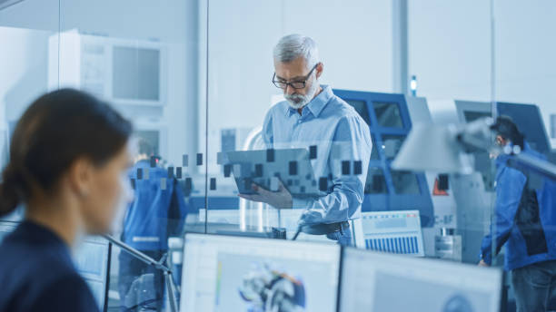 istock Modern Factory: Senior Engineer Looks at Camera, Holding Laptop, Inspecting, Overlooking Workshop Operation. Manufactory Has Professionals Working on CNC Machinery, Robot Arm. 1219894402