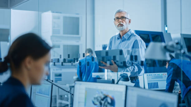 istock Modern Factory: Senior Engineer Looks at Camera, Holding Laptop, Inspecting, Overlooking Workshop Operation. Manufactory Has Professionals Working on CNC Machinery, Robot Arm. 1219894458