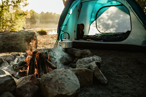 Campsite with a tent, kettle and burning fire pit in countryside. Camping by a lake in forest.