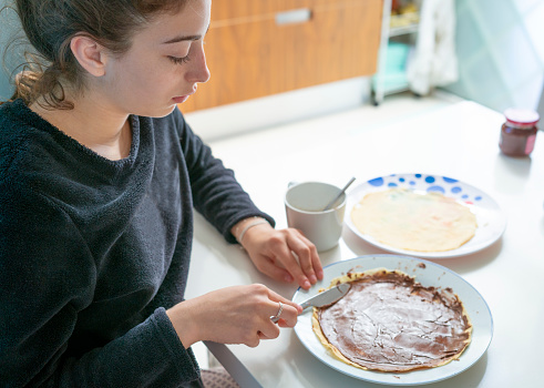 Teenager girl spreading chocolate cream for breakfast on a Crepe in a white table