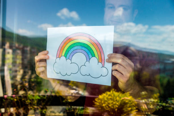 istock Man holding rainbow in window during Covid-19 pandemic 1219903193