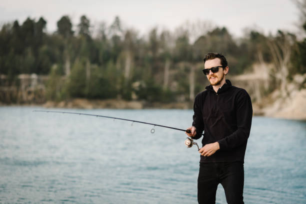 istock Fisherman with rod, spinning reel on river bank. Sunrise. Man catching fish, pulling rod while fishing from lake or pond. Fishing for pike, perch, carp on beach lake or pond. Background wild nature. 1219911372