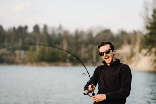 istock Fisherman with rod, spinning reel on river bank. Sunrise. Man catching fish, pulling rod while fishing from lake or pond. Fishing for pike, perch, carp on beach lake or pond. Background wild nature. 1219911407