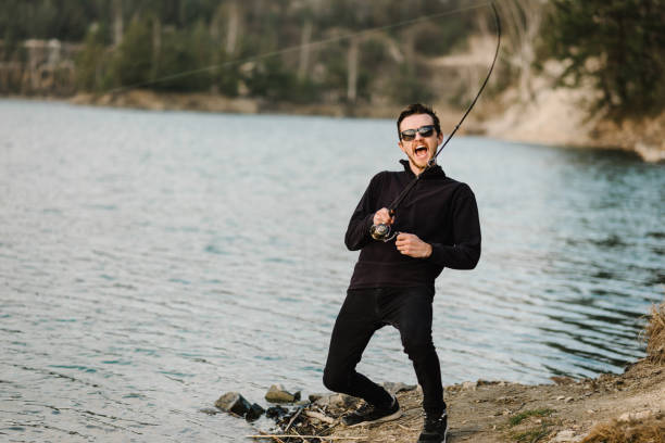istock Fisherman with rod, spinning reel on the river bank. Man catching fish, pulling rod while fishing from lake or pond with text space at weekend. Fishing for pike, perch, carp on beach lake or pond. 1219911413