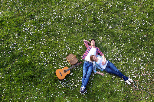 Happy heterosexual couple enjoying picnic time lying on lawn with an acoustic guitar. Both Caucasian in casual clothing, about 20-25 years old.