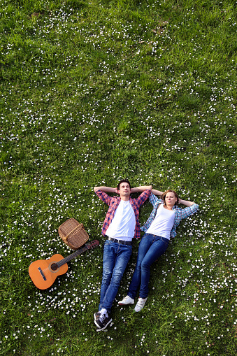 Happy heterosexual couple enjoying picnic time lying on lawn with an acoustic guitar. Both Caucasian in casual clothing, about 20-25 years old.