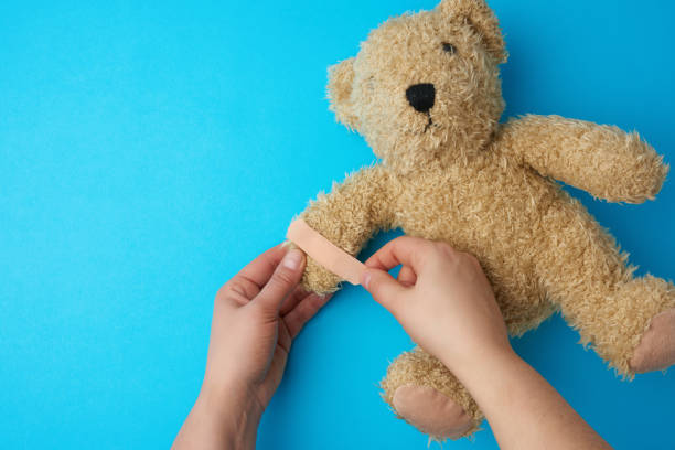 two hands holding a teddy bear and gluing an adhesive plaster on a paw two hands holding a teddy bear and gluing an adhesive plaster on a paw, blue background, top view behavior teddy bear doll old stock pictures, royalty-free photos & images