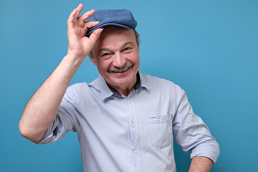 Cheerful senior man greets by taking off a hat. Studio shot on blue wall.