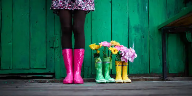 Legs of young woman in black tights, skirt with floral print and pink rubberboots standing against green painted wall or fence outside