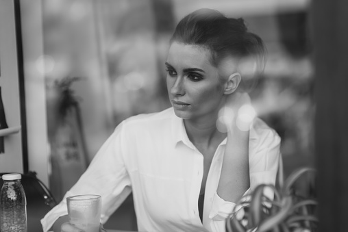 A beautiful young girl sits in a cafe behind a glass with a cup of coffee and looks thoughtfully out the window. Black and white art photo through glass with reflection. Soft selective focus.