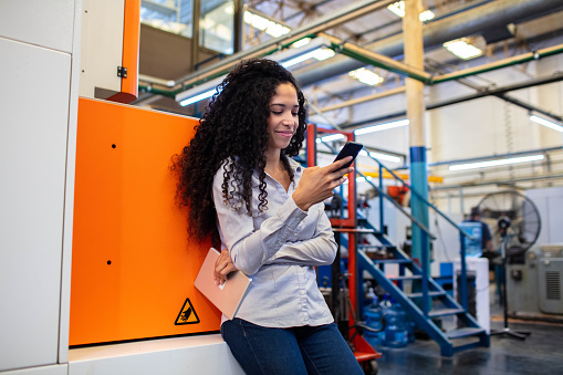 Young female industrial worker using her mobile phone while leaning to a machine in factory. Woman engineer in company shop floor looking at the cell phone.