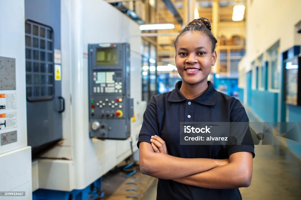 Woman technician standing in factory shop floor Portrait of a young woman technician standing in factory shop floor. Female in uniform looking at camera with her hands folded. Manufacturing Stock Photo