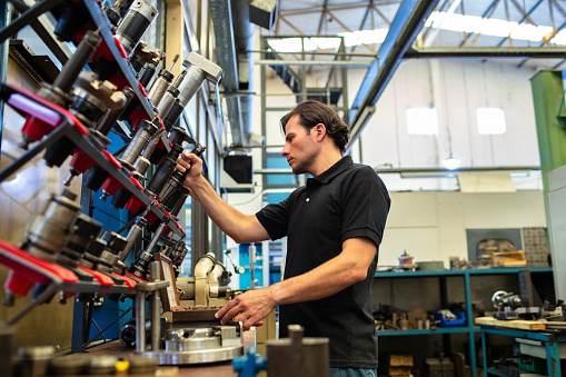 Male worker working in manufacturing plant. Man in uniform picking up a tool from rack in factory.