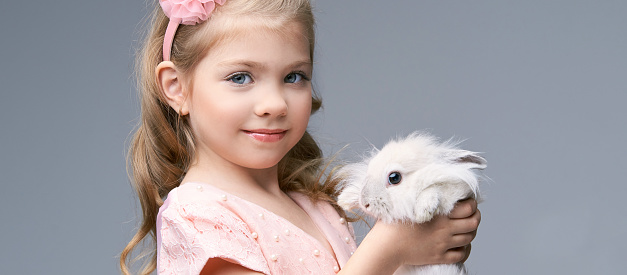 Little pretty girl holding fur rabbit baby. White cute pet at studio. Happy easter hug. Female person hand. Smile people child. Young people.