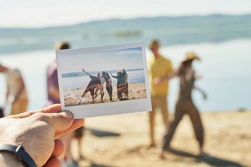 Close-up view of male hand holding polaroid photo print with young people hanging out on lake on beautiful summer day