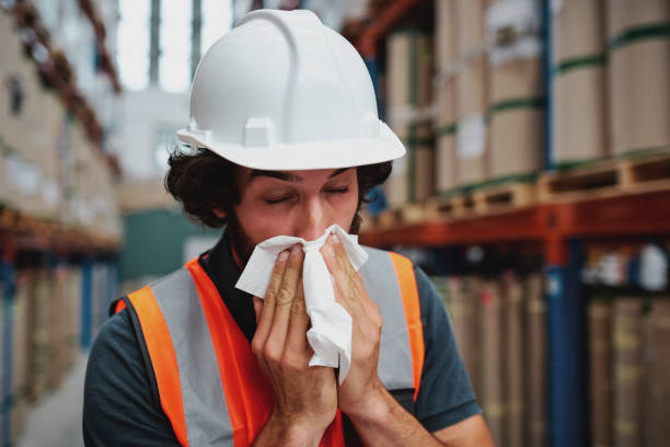 istock Young warehouse worker feeling sick while sneezing and coughing at factory standing near packed cardboard boxes on shelf 1219943064