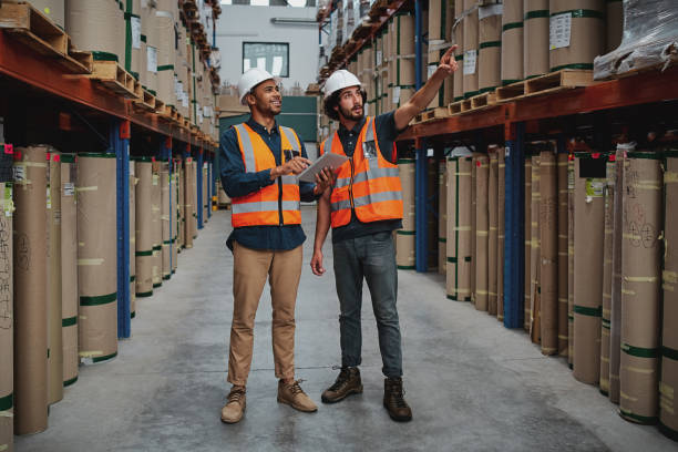 istock Full length of joyful professional managers working together while being in the warehouse holding digital tablet pointing towards cardboard box on shelf 1219943145