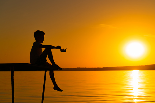 In the evening a small boy sits on the bridge over the calm river with a paper boat. A child is watching a beautiful sunset in summer . Origami.