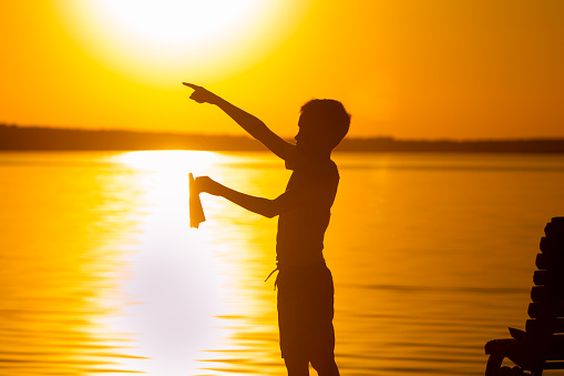 a little child stands on the background of the lake at sunset. In the left hand he holds a paper airplane, and with the right hand he points with his finger into the distance