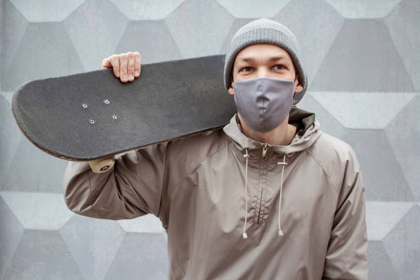 europäischer kerl in medizinischer maske hält skateboard auf seiner schulter. grauer beton wand hintergrund. skateboarder in grauer jacke und hut. coronavirus-schutzmaßnahmen. - isolated cold virus head and shoulders cold and flu stock-fotos und bilder