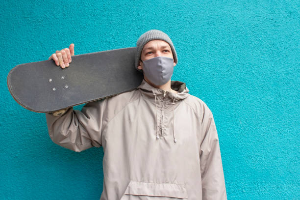 europäischer kerl in medizinischer maske hält skateboard auf seiner schulter. gesättigte türkis enk. skateboarder in grauer jacke und hut. coronavirus-schutzmaßnahmen. - isolated cold virus head and shoulders cold and flu stock-fotos und bilder