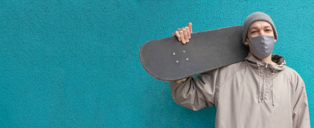 istock European guy in face mask holds skateboard on shoulder. Turquoise background. Horizontal banner for web site or social networks. Space for text. Skateboarder in a hat. Coronavirus protection measures. 1219945421