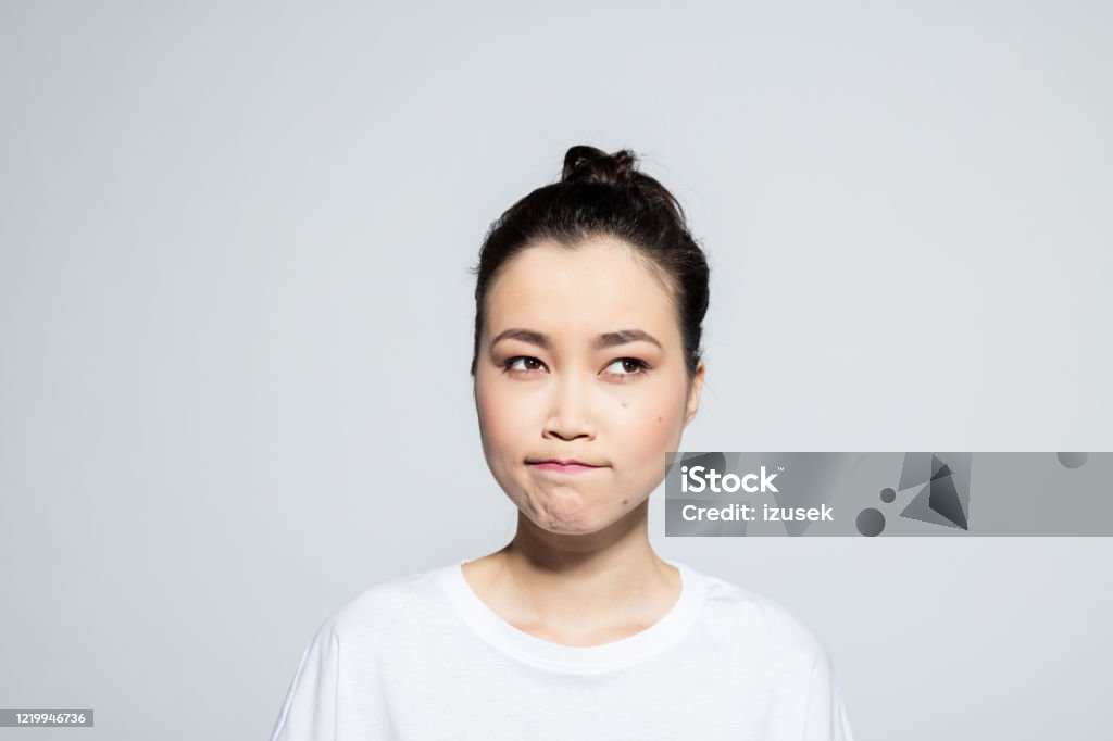 Disappointed asian young woman Headshot of beautiful asian young woman wearing white t-shirt, looking away and grimacing. Studio shot, grey background. Human Face Stock Photo